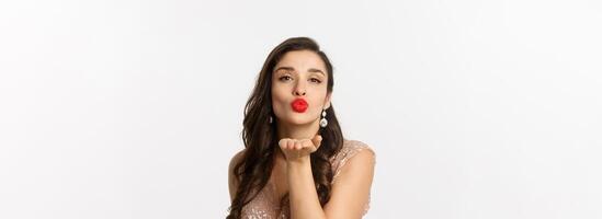 Concept of New Year celebration and winter holidays. Close-up of sensual young woman in dress, pucker lips and blowing air kiss at camera, white background photo