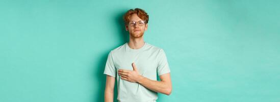 Proud young redhead man in glasses holding hand on heart, listening national anthem with respect, standing in t-shirt against turquoise background photo