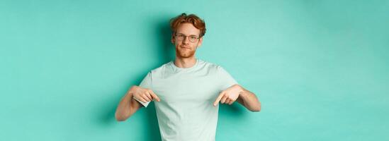 Skeptical and bothered man with ginger hair and beard, wearing glasses and t-shirt, smirk and point fingers down, showing promo with judgy face, turquoise background photo