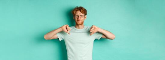 Disappointed caucasian guy with red messy hair and glasses showing thumbs down, dislike something, frowning upset and displeased, standing over mint background photo