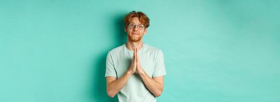Cute redhead man asking for help, saying please with namaste gesture, pleading or begging for favour, looking silly at camera, standing over mint background photo