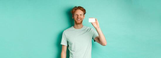Shopping concept. Handsome redhead man in t-shirt showing plastic credit card and smiling, standing over turquoise background photo