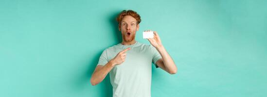 Shopping concept. Handsome redhead man in t-shirt showing plastic credit card and smiling, standing over turquoise background photo