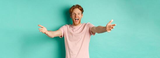 Friendly and happy young man with ginger hair, stretch out hands in warm welcome, reaching for hug and smiling joyfully, standing in t-shirt over mint background photo