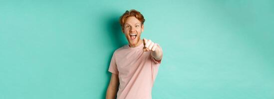Excited young man with red hair checking out something cool, pointing finger at camera and smiling fascinated, standing over turquoise background photo