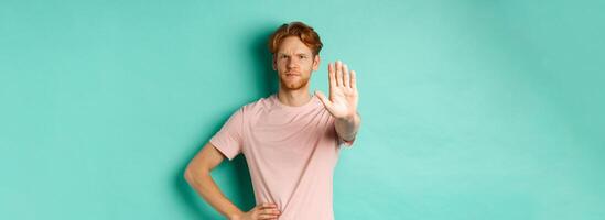 Serious young man with red hair and beard telling to stop, showing palm and frowning, disapprove and prohibit something bad, standing over turquoise background photo