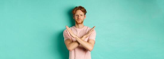 Skeptical redhead man in t-shirt saying no, cross arms on chest and frowning disappointed, prohibit something bad, standing over turquoise background photo