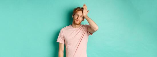 Annoyed and bothered redhead male model showing facepalm gesture, standing over mint background photo