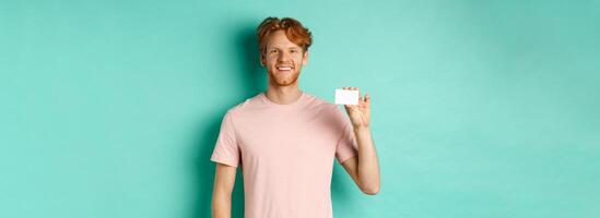 Handsome young man smiling and showing plastic credit card, standing in t-shirt against turquoise background photo