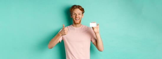 Cheerful male bank client in t-shirt showing thumb up and plastic credit card, smiling satisfied at camera, standing over turquoise background photo