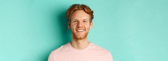 Close up of redhead bearded guy in pink t-shirt, smiling with white perfect teeth and looking at camera, standing over turquoise background photo