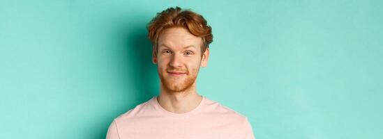Close up of redhead bearded man looking pleased, nod in approval and smiling, standing in pink t-shirt against turquoise background photo