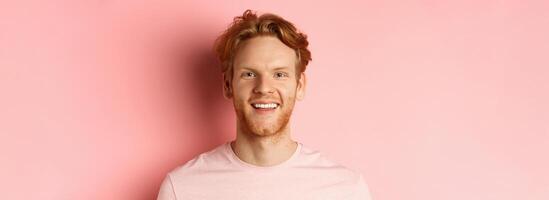 Headshot of happy redhead man with beard and white teeth, smiling excited at camera, standing over pink background photo
