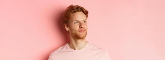 Handsome european male model with red hair and beard, turn head and looking pleased at copy space on left side, standing over pink background photo