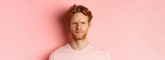 Close up of suspicious redhead guy frowning, staring at camera doubtful, express disapproval, standing over pink background photo