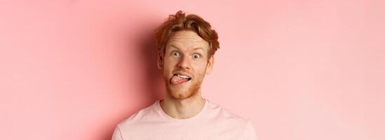 Headshot of funny redhead guy showing tongue, making silly faces at camera, standing joyful against pink background photo