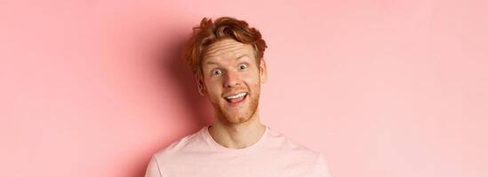Headshot of funny redhead guy showing tongue, making silly faces at camera, standing joyful against pink background photo