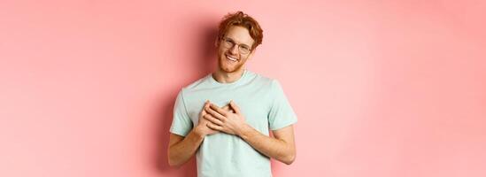 Attractive young man with ginger hair, holding hands on heart and smiling grateful, saying thank you, express gratitude, standing over pink background photo