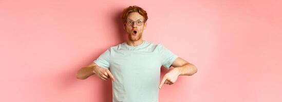 Image of amazed young man with red hair and beard, wearing glasses and t-shirt, pointing fingers down and staring excited at camera, pink background photo