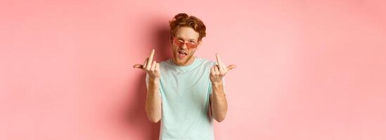 Arrogant and careless redhead man in sunglasses dont give a fuck, showing middle fingers at camera and frowning, standing over pink background photo