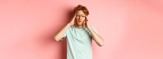 Portrait of redhead man in crooked glasses touching head and feeling dizzy or nauseous, having hangover or headache, standing over pink background photo