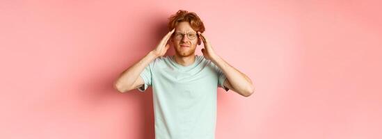 Portrait of redhead man in crooked glasses touching head and feeling dizzy or nauseous, having hangover or headache, standing over pink background photo