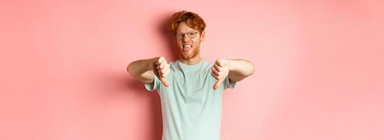 Disgusted redhead man in t-shirt and glasses showing thumbs down and tongue, grimacing from aversion, standing over pink background photo