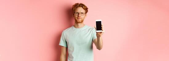 Disappointed male model frowning, showing smartphone screen, standing over pink background photo
