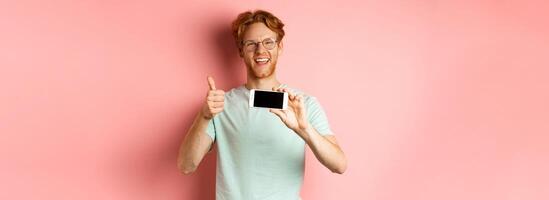 Happy european man with ginger hair and glasses, showing mobile blank screen horizontally and thumbs-up, recommend online promo, pink background photo