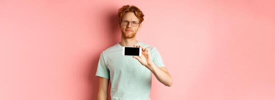 Skeptical redhead guy in glasses showing smartphone screen horizontally, smirk and frowning disappointed, standing over pink background photo