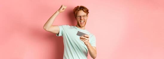 Happy redhead man winning in mobile game, raising hand up and shouting yes with joy, celebrating victory, looking at smartphone, standing over pink background photo