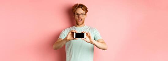 Amazed redhead man gasping and staring with awe phone, showing blank smartphone screen horizontally, standing over pink background photo