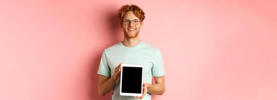 Funny redhead guy in glasses demonstrate digital tablet screen, smiling at camera, standing over pink background photo