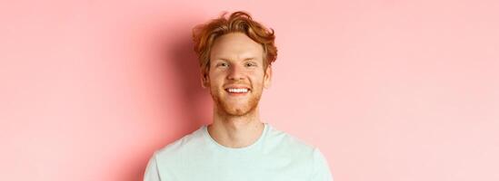 Happy young man with beard and messsy red haircut, smiling with white teeth and cheerful expression, standing over pink background photo