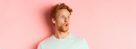 Headshot portrait of surprised redhead man with beard, looking left and saying wow, raising eyebrows amazed, standing over pink background photo