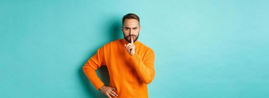 Angry bearded guy telling be quiet, hushing and frowning displeased, shut up gesture, standing over turquoise background photo