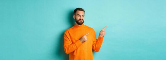 Displeased and skeptical guy grimacing, showing bad product, pointing fingers left with disappointment, standing turquoise background photo