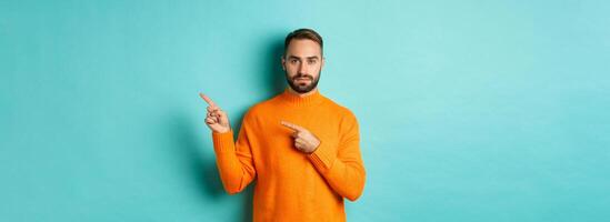 Handsome and serious bearded man in orange sweater pointing right, showing advertisement or logo, standing over turquoise background photo