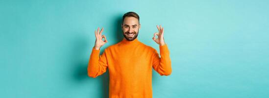 Image of confident smiling man showing okay sign, approve and agree, guarantee quality, standing over light blue background photo