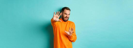 Disgusted man cringe and extending hands in protection, begging for mercy, looking at something with aversion, standing over light blue background photo