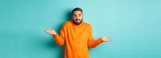 Confused caucasian man with beard, shrugging and looking clueless, standing indecisive against turquoise background photo