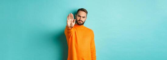 Serious frowning man showing stop gesture, telling no, prohibit and disagree, standing over light blue background photo