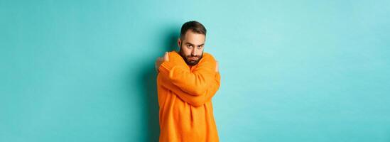 Timid man feeling offended and defensive, hugging himself and looking suspicious at camera, standing over light blue background photo