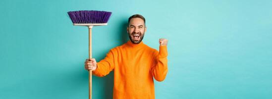 Image of encouraged and motivated man getting ready for cleaning, holding broom and making fist pump gesture, standing over turquoise background photo