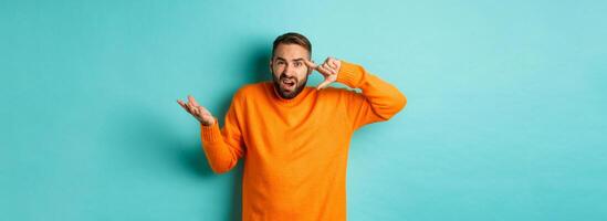 Angry man pointing at head, are you crazy gesture, scolding someone, standing over light blue background photo