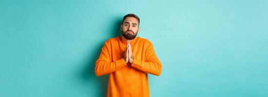 Sulking man begging for help, asking for favour, holding hands in pray and pouting, standing over turquoise background photo