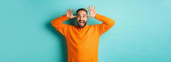 Image of handsome caucasian man making funny faces, mocking someone and smiling, standing against light blue background photo