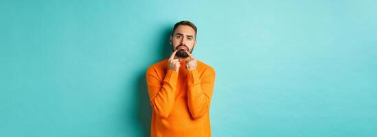 Image of gloomy bearded man, making sad face and frowning, standing upset in orange sweater against turquoise background photo