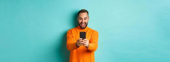 Image of man reading awesome message on mobile phone, looking at smartphone screen with amazement, standing over light blue background photo
