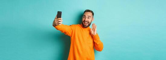 Happy young man calling, talking online with mobile phone, saying hello to smartphone camera and waving hand friendly, standing over light blue background photo